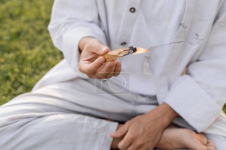 partial view of man in white cotton clothes holding burning palo santo stick while meditating outdoors