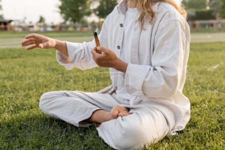Téléchargez les photos : Vue partielle de l'homme en vêtements blancs tenant palo santo fumant bâton et méditant dans la pose facile sur la pelouse verte - en image libre de droit