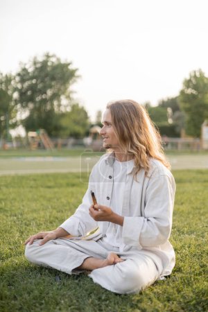 Unbekümmerter Mann in weißer Baumwollkleidung meditiert in lockerer Pose mit duftendem Palo Santo Stick auf grünem Gras im Freien