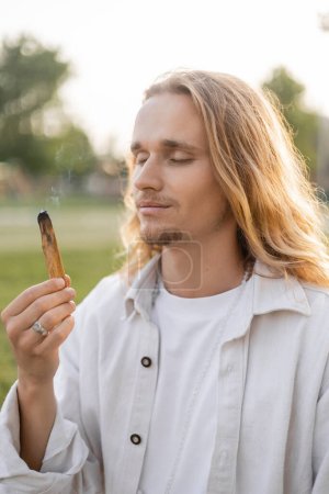 Téléchargez les photos : Homme souriant avec les cheveux longs et les yeux fermés appréciant la saveur de palo santo fumant bâton à l'extérieur - en image libre de droit