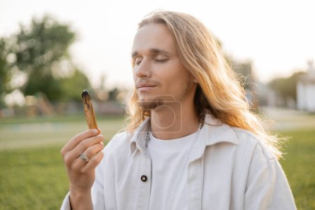 Téléchargez les photos : Heureux homme aux cheveux longs avec les yeux fermés tenant palo santo fumant bâton tout en méditant à l'extérieur - en image libre de droit