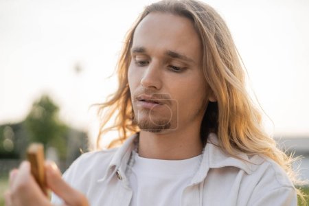 portrait of young man with long fair hair holding blurred palo santo stick outdoors