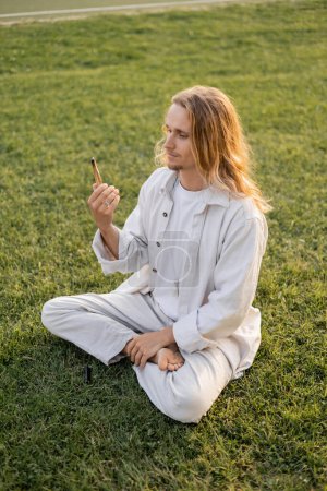 young yoga man in white clothes looking at fragrant palo santo stick while sitting in sukhasana pose on green grass