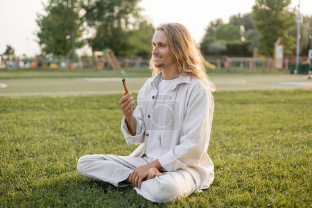 feliz hombre de pelo largo sosteniendo palo santo aromático mientras medita en pose fácil sobre césped verde
