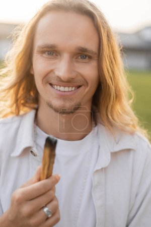 Téléchargez les photos : Joyeux homme de yoga aux cheveux longs tenant palo santo fumant bâton et souriant à la caméra à l'extérieur - en image libre de droit