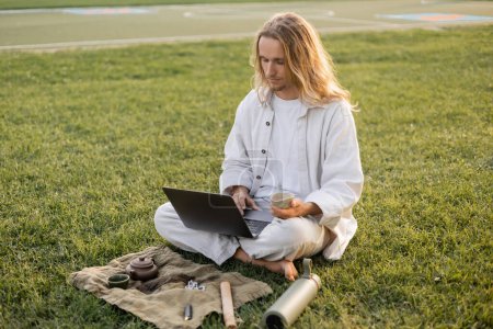 young man in white clothes holding puer tea and using laptop while sitting near thermos and ceramic teapot on green lawn