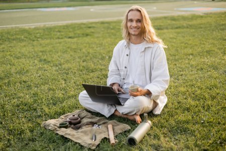 Photo for Happy long haired man sitting with laptop and clay cup near thermos and linen rug with teapot on grassy lawn - Royalty Free Image