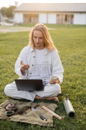 Langhaariger Mann in Leinenkleidung mit Videoanruf am Laptop, während er mit purem Tee in der Nähe von Teppich mit Tonbecher und Teekanne auf grünem Rasen sitzt