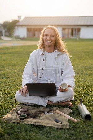 hombre despreocupado sonriendo a la cámara mientras está sentado en la hierba verde con el ordenador portátil y taza de arcilla cerca de termos y alfombra de lino con tetera