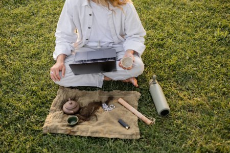 vista parcial del hombre del yoga sentado en el césped verde con el ordenador portátil y el té puro cerca del termo y la alfombra de lino con la tetera de arcilla y cuentas de mala