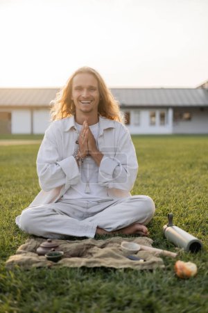 Unbekümmerter Mann in Leinenkleidung meditiert neben Tontassen und Teekanne bei Teezeremonie auf grünem Rasen im Grünen