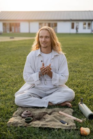 Téléchargez les photos : Homme aux cheveux longs souriants en vêtements blancs assis près du tapis de lin avec théière en céramique et regardant loin dans la campagne - en image libre de droit