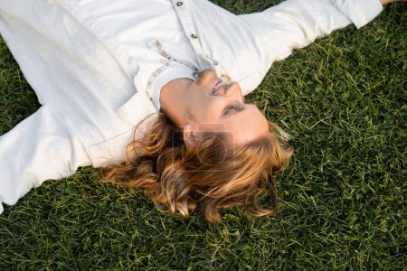 top view of carefree long haired man in white shirt lying and smiling on green lawn
