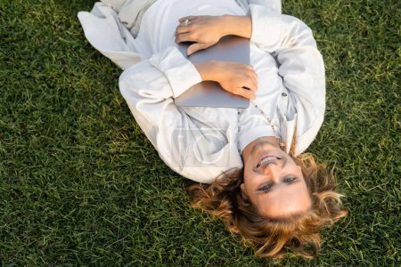 Téléchargez les photos : Vue de dessus de l'homme insouciant en blanc vêtements élégants couché avec ordinateur portable sur la pelouse verte - en image libre de droit