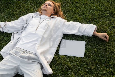 high angle view of young yoga man in white clothes lying near laptop on green field 