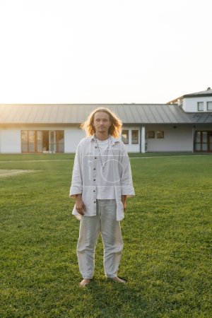 full length of yoga man in cotton clothes standing on grassy lawn near cottage on background