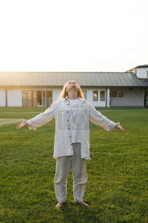 Photo for Full length of man in linen clothes meditating with outstretched hands and raised head on green lawn near countryside cottage - Royalty Free Image