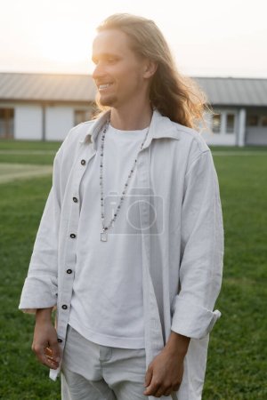 joven hombre de yoga de pelo largo en camisa de lino y perlas sonriendo mientras está de pie en el campo al aire libre