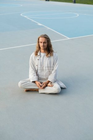 Téléchargez les photos : Longueur complète de l'homme élégant avec de longs cheveux blonds méditant dans la pose de yoga facile sur le stade à l'extérieur - en image libre de droit