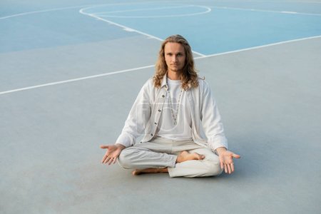 Foto de Longitud completa del hombre de pelo largo en ropa blanca meditando en pose fácil mientras está sentado en el estadio al aire libre - Imagen libre de derechos