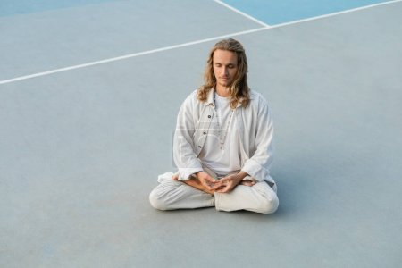 Téléchargez les photos : Homme aux cheveux longs en vêtements de coton blanc pratiquant le yoga en pose de lotus sur le stade extérieur - en image libre de droit