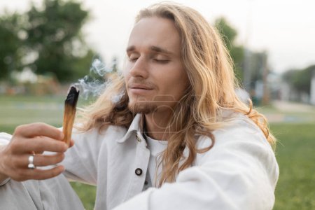 Téléchargez les photos : Homme heureux avec les yeux fermés tenant palo santo fumant bâton pendant rituel spirituel à l'extérieur - en image libre de droit