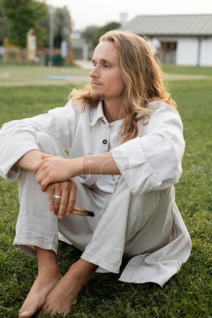 calm barefoot man in white linen clothes looking away while sitting with aromatic palo santo stick outdoors