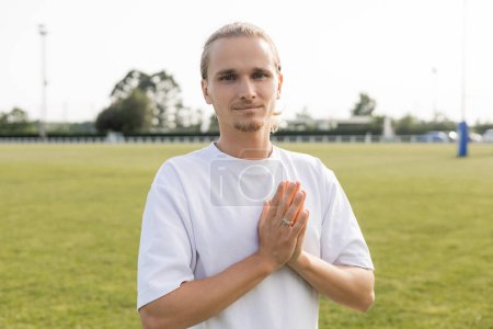 young and positive man in white t-shirt showing anjali mudra gesture and looking at camera on blurred outdoor stadium