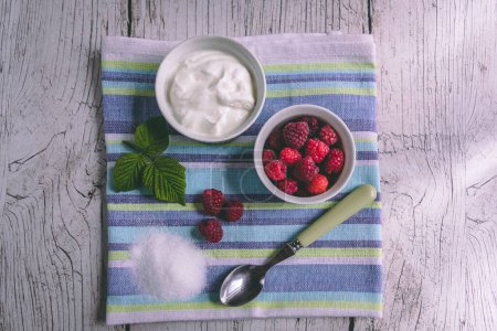 Foto de Una toma de vista superior de un tazón pequeño de frambuesas, yogur blanco y menta en una toalla de rayas azules - Imagen libre de derechos