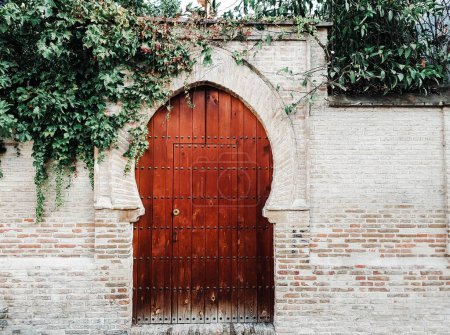 Photo for A closeup of a wooden arched Moroccan style door with stone high fence around and espaliered plant on - Royalty Free Image