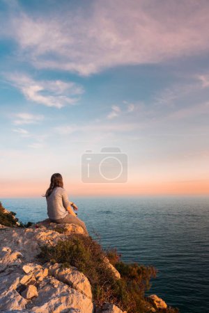 Photo for Young woman from behind watching the sea and resting on the edge of a cliff during sunset - Royalty Free Image