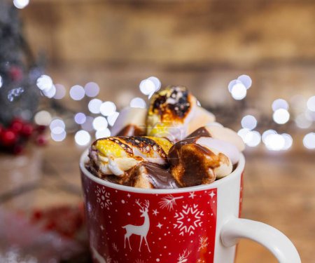 Foto de El sabroso chocolate caliente con malvaviscos en una taza roja de Navidad sobre el fondo borroso de las luces - Imagen libre de derechos