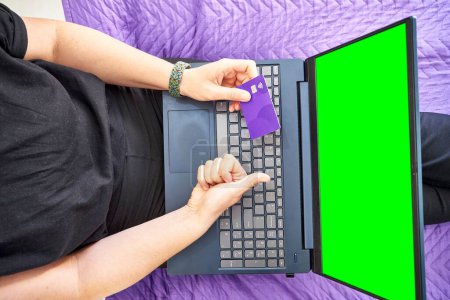 Téléchargez les photos : Une vue de dessus des mains d'une personne pointant vers l'écran vert d'un ordinateur portable et tenant une carte bancaire - en image libre de droit