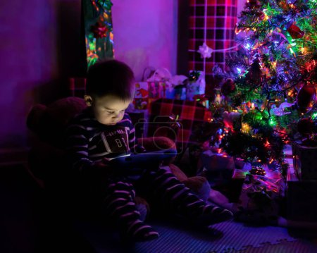 Téléchargez les photos : Un jeune garçon et tout-petit assis dans l'obscurité à côté du sapin de Noël et présente jouer sur la tablette - en image libre de droit