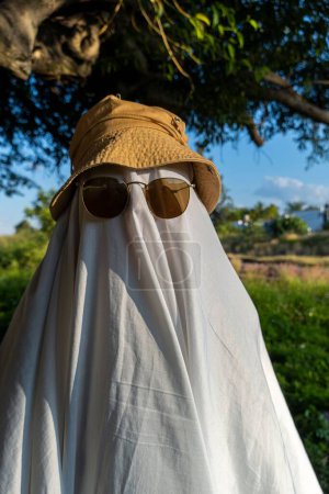 Photo for A man dressed as a ghost from a sheet and sunglasses crosses the road along the crosswalk - Royalty Free Image
