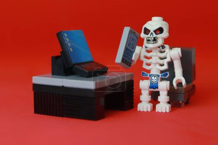 Téléchargez les photos : Plan rapproché d'un squelette de jouet avec des yeux rouges, travaillant de la maison et tenant un téléphone, debout à côté d'un bureau avec un ordinateur dessus - en image libre de droit
