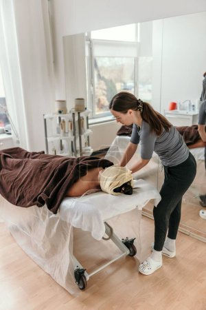 Téléchargez les photos : Masseuse faisant anti stress massage du corps sur la femme allongée sur une table de massage enveloppée dans une serviette - en image libre de droit