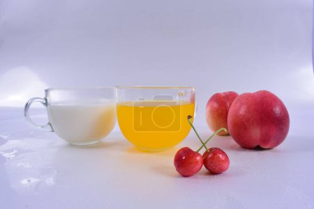 Téléchargez les photos : Un gros plan de deux verres avec du jus d'orange et du lait près des pêches et des cerises sur une surface blanche - en image libre de droit