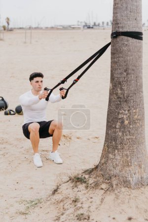 Téléchargez les photos : Un jeune homme sportif espagnol en forme faisant des exercices de traction d'épaule avec une corde à la plage - en image libre de droit