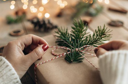 Foto de Foto de cerca de la mano femenina con esmalte de uñas rojo, envoltura o apertura regalo de Navidad envuelto en papel marrón y decorado con ramita de abeto verde. - Imagen libre de derechos