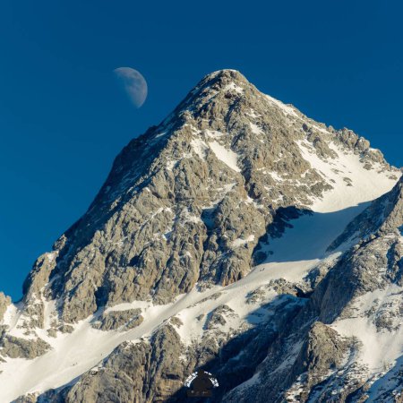 Photo for A low angle shot of a rocky mountain peak in snow and the half moon in the blue sky - Royalty Free Image