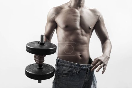 Photo for The concept of weight loss, a muscular shirtless man holding a dumbbell and wearing oversize jeans - Royalty Free Image