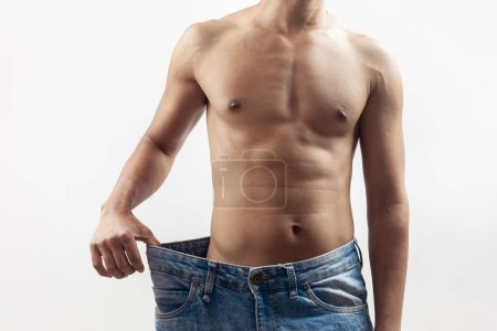 Photo for The concept of weight loss, a fit young man wearing a loose blue jeans showing his weight loss progress - Royalty Free Image