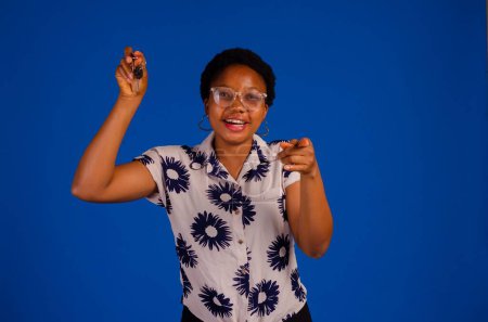 Foto de Una mujer africana sosteniendo las llaves y apuntando a la cámara aislada sobre un fondo azul. - Imagen libre de derechos