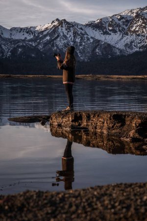 Téléchargez les photos : Plan vertical d'une jeune femme debout au bord d'un lac et photographiant la montagne enneigée des hautes terres - en image libre de droit