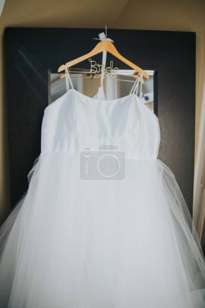 Téléchargez les photos : Une verticale de la robe blanche de la mariée accrochée à un cintre devant un miroir - en image libre de droit