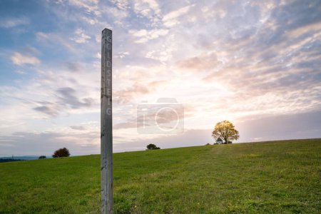Téléchargez les photos : Une vue coucher de soleil avec un pilier en bois isolé dans un champ avec une chapelle sous un arbre en arrière-plan - en image libre de droit