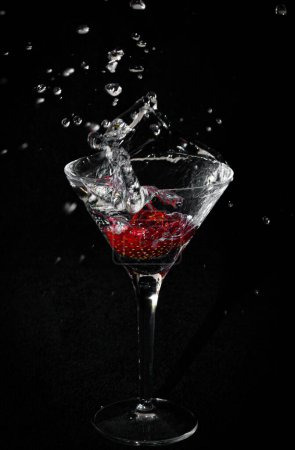 A vertical shot of a glass of cocktail with small red berries and splashing water on black background