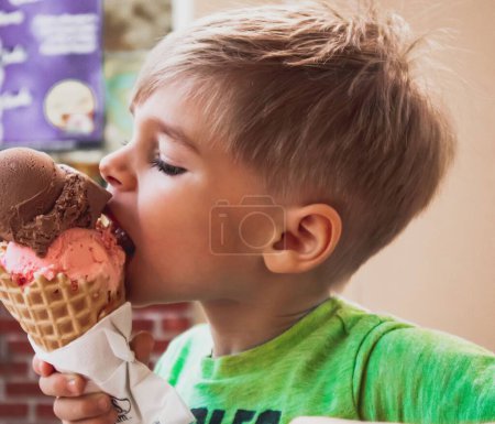 Téléchargez les photos : Un gros plan d'un jeune garçon mangeant un cône de crème glacée multi-cuillère à l'extérieur fond flou - en image libre de droit