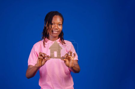 Photo for A portrait of a young black woman holding a paper home for a real estate concept on a blue background. - Royalty Free Image